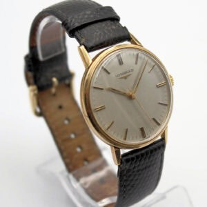 A Longines solid 9ct Gold Presentation Watch from 1977, featuring a round silver dial with gold hour markers and a black textured leather strap, displayed on a clear watch stand.