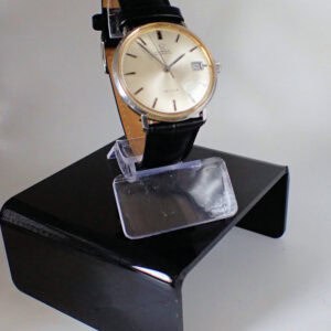 The Omega De Ville Date Automatic features a round silver dial, gold case, black leather strap, and date display, displayed on a clear and black stand against a plain background.