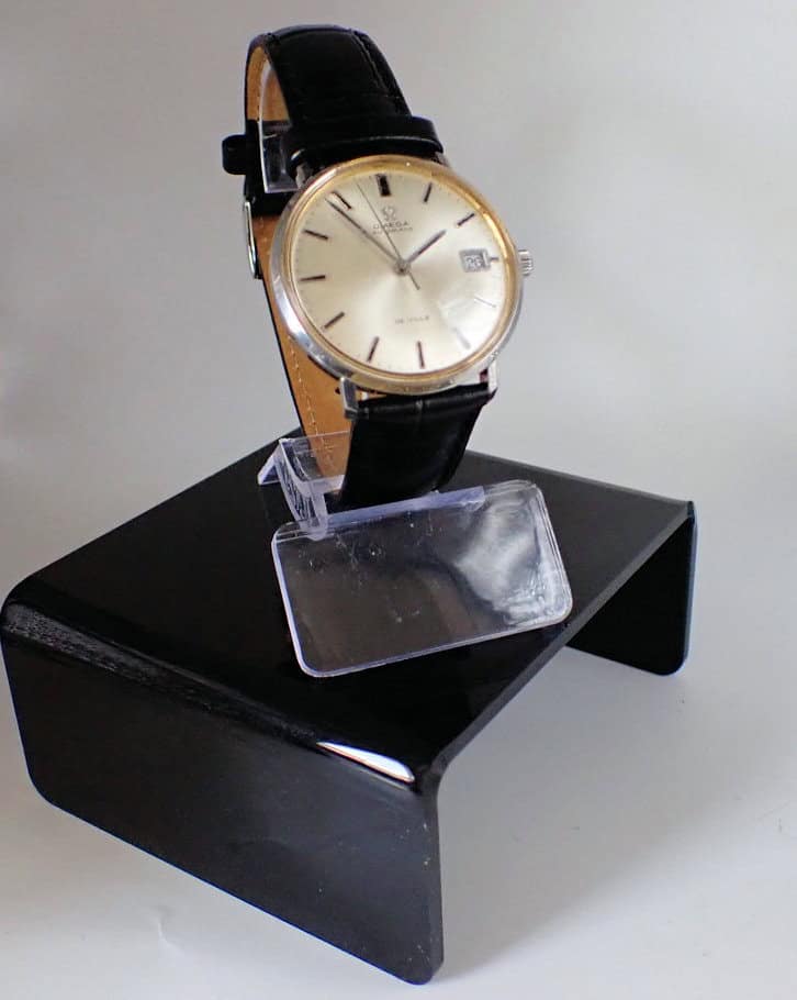 The Omega De Ville Date Automatic features a round silver dial, gold case, black leather strap, and date display, displayed on a clear and black stand against a plain background.