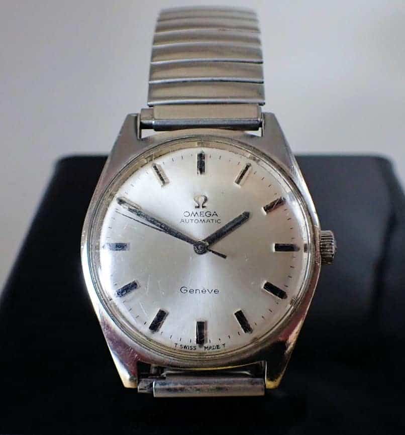 The Omega Genève Automatic Wristwatch features a silver dial, minimalist hour markers, and a metal link strap, beautifully presented against a dark background.