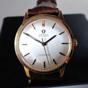 An Omega Seamaster Automatic Circa 1960s wristwatch with a gold case, white dial, gold hour markers, and brown leather strap, displayed upright on a black stand.