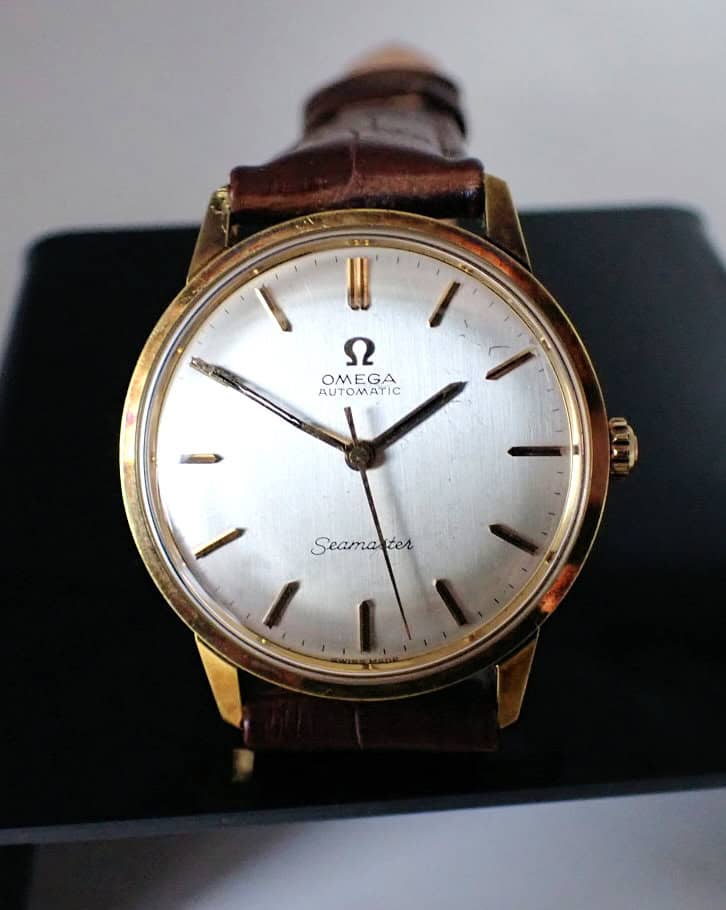 An Omega Seamaster Automatic Circa 1960s wristwatch with a gold case, white dial, gold hour markers, and brown leather strap, displayed upright on a black stand.