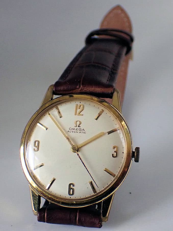 The OMEGA Automatic Gold Plated Watch 1961 features a round gold case, white dial, gold hour markers, and a brown leather strap, displaying the time as 4:16.