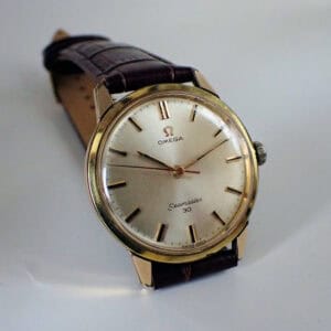 An Omega Seamaster 30 Manual Wind 1963 wristwatch with a gold case, leather strap, and silver dial with gold hour markers and hands, shown on a light background.