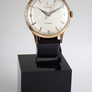 A vintage Lanco 17 Jewel Incabloc wristwatch in a 9ct solid gold case with a white dial, black leather strap, and silver hour markers is displayed on a black stand. "INCABLOC" appears on the dial.