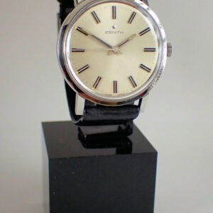 The Zenith Stainless Steel Presentation Watch 1969, featuring a round white dial, silver case, black leather strap, and black hour markers, is displayed on a black stand with its original box and dated guarantee.