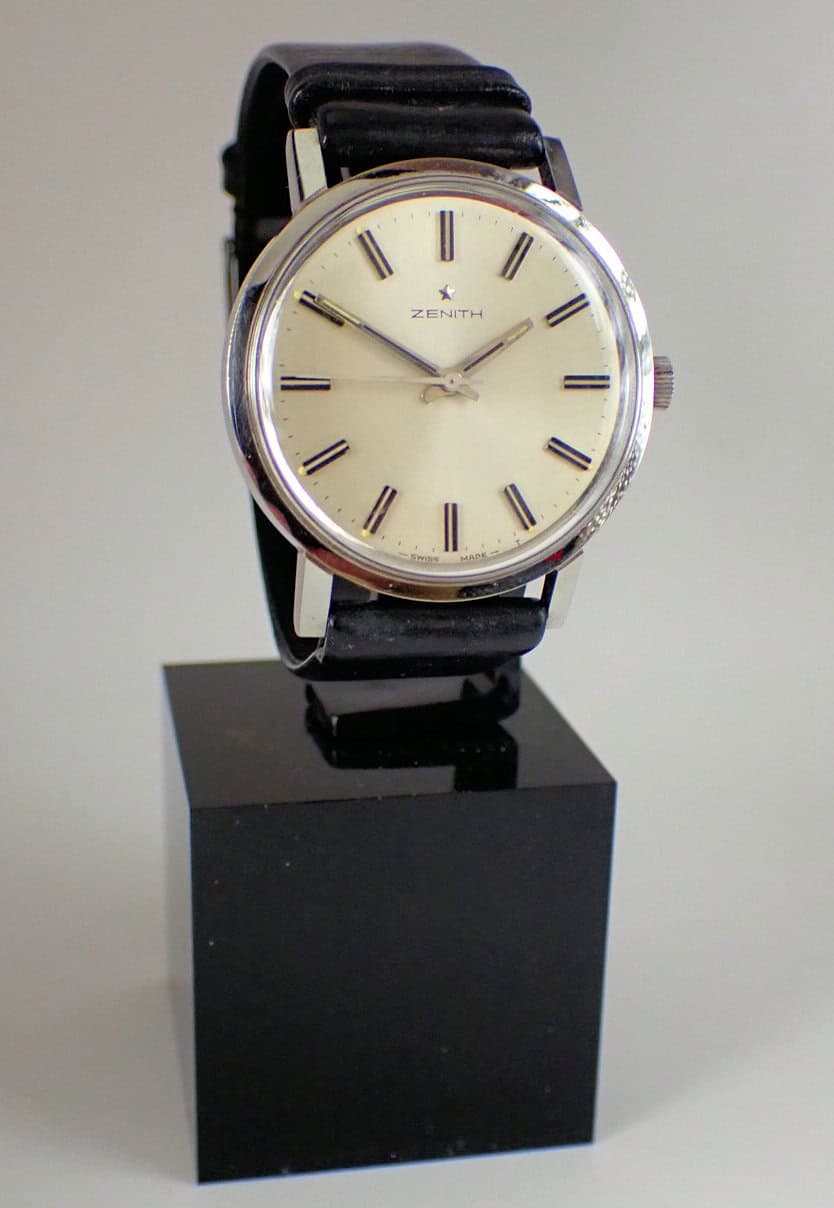 The Zenith Stainless Steel Presentation Watch 1969, featuring a round white dial, silver case, black leather strap, and black hour markers, is displayed on a black stand with its original box and dated guarantee.