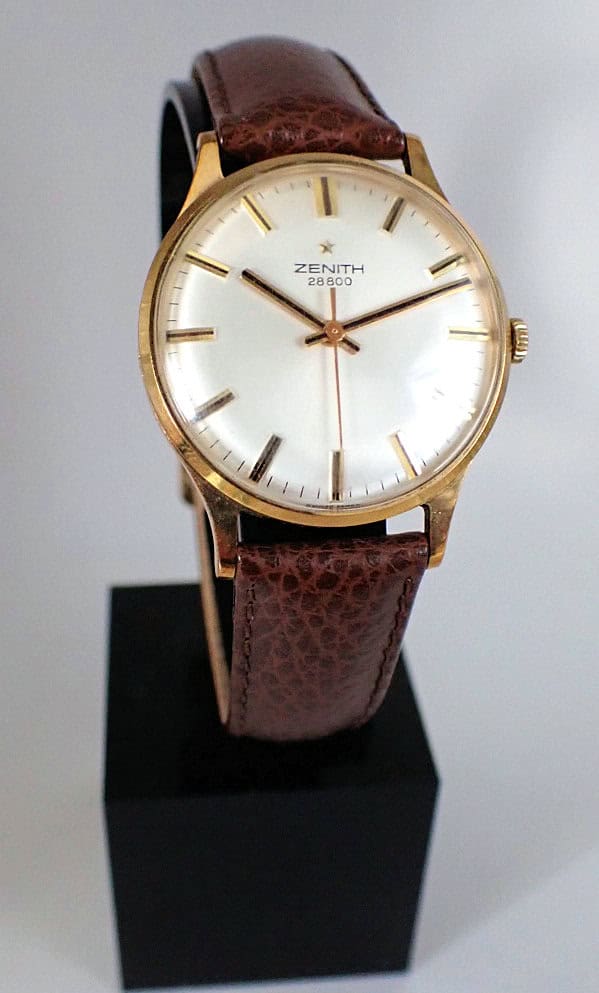 A Zenith 28800 Mechanical Solid 9ct Gold Watch with a white dial, gold hour markers, and brown leather strap is displayed on a black stand against a plain background.