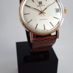 The Zenith Date Automatic Solid 9ct Gold Presentation Watch features a round white dial, gold case, brown textured leather strap, and date display at 3 o'clock, displayed on a black stand against a white background.