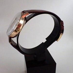 Leather strap watch with a gold-tone case and clear dial, displayed on a black stand.
