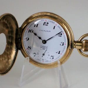The Excalibur Half Hunter Quality 1970s Watch, a gold pocket watch with its cover open to show a white face, black numerals, and a small seconds dial, is displayed on a clear stand with its ornate winding crown visible.