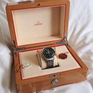 The Omega Speedmaster Professional 'Moon Watch' 2009 with papers, featuring a black dial and silver case, is shown in an open wooden Omega box with cream interior, accompanied by a red wax seal tag and a small white tag on a gold string.