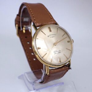 The Rotary Incabloc Mechanical 17 Jewels Vintage Wristwatch features a gold case, cream dial reading “Rotary” and “17 Jewels Incabloc,” plus a brown leather strap, displayed on a clear stand.