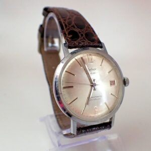 A Hafner Superautomatic Incabloc Swiss Made vintage watch with a round silver dial, simple hour markers, date window at “29”, and a brown textured leather strap, displayed on a clear stand against a light background.
