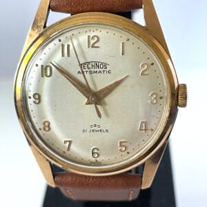A vintage Technos Automatic 21 Jewels 1960s wristwatch featuring a gold case, brown leather strap, white dial with gold numerals and hands, TECHNOS AUTOMATIC 21 JEWELS text, and a logo above SWISS.