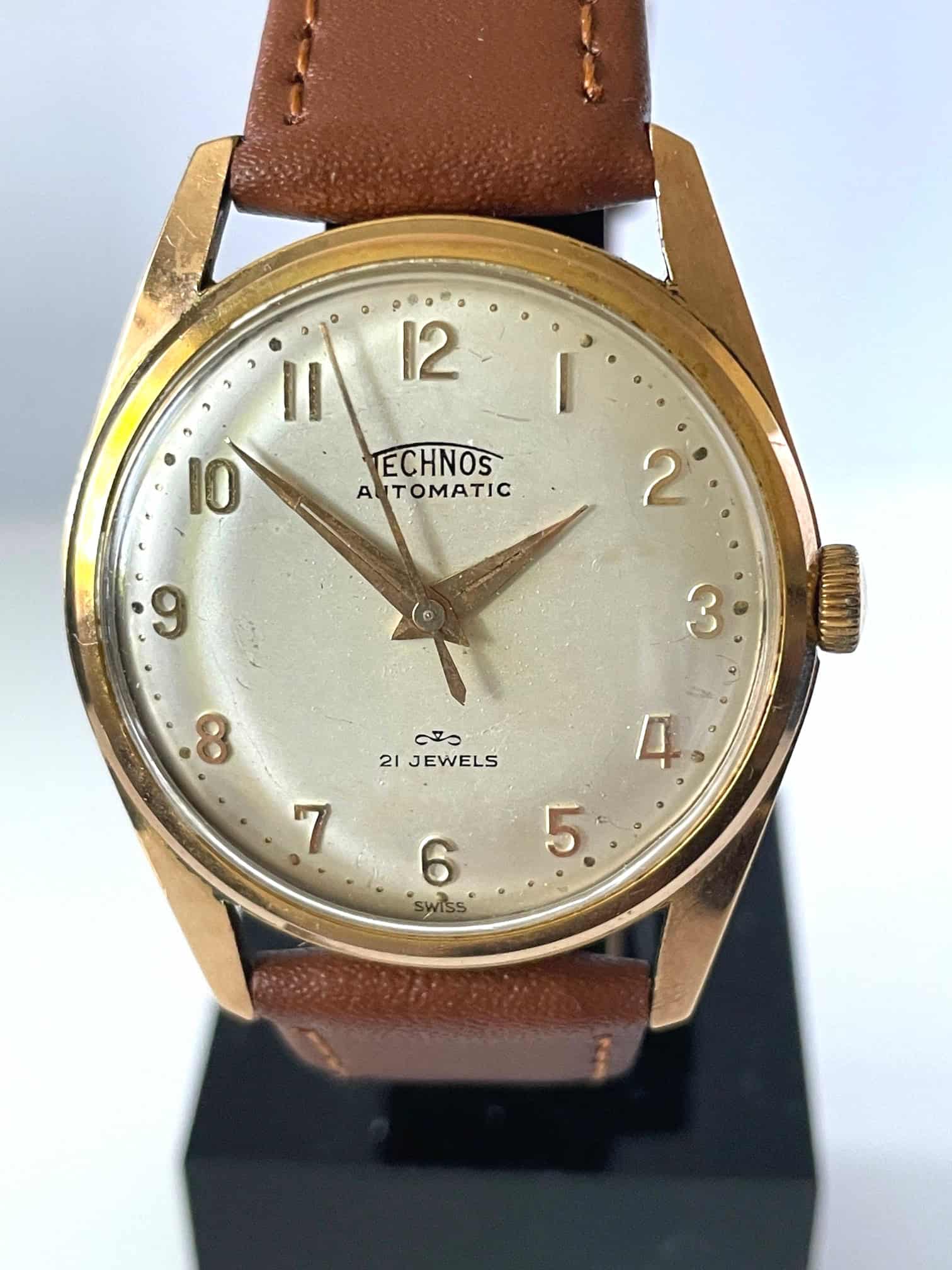 A vintage Technos Automatic 21 Jewels 1960s wristwatch featuring a gold case, brown leather strap, white dial with gold numerals and hands, TECHNOS AUTOMATIC 21 JEWELS text, and a logo above SWISS.