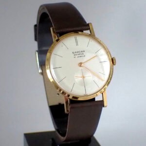 A vintage Sarcar Genève 17 Jewels Mechanical wristwatch from circa 1970 featuring a white dial with black markers, gold hands, and a brown leather strap, displayed upright on a black stand.