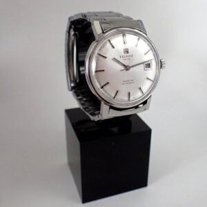 The Tissot Seastar Automatic 1970s wristwatch features a silver metal strap, round white face, and date display, shown upright on a black rectangular plinth against a plain background.