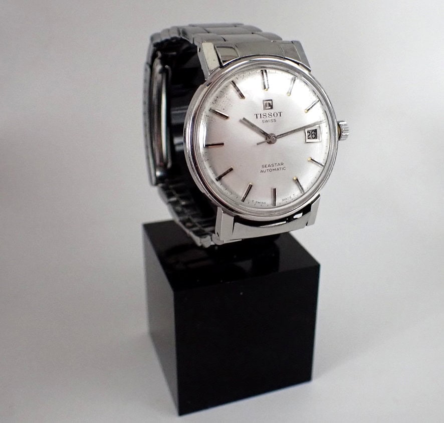 The Tissot Seastar Automatic 1970s wristwatch features a silver metal strap, round white face, and date display, shown upright on a black rectangular plinth against a plain background.