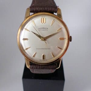 A rare Derrick 9ct Gold Automatic gents' watch featuring a gold-tone case, brown leather strap, white dial with gold markers and hands, and DERrick AUTOMATIC SHOCK PROTECTED inscribed on the face.