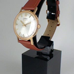 An Oris 17 Jewel Mechanical wristwatch with a brown leather strap, gold case, and white dial is displayed upright on a black stand against a plain background.