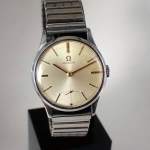 A vintage Omega (Seamaster) mechanical 1964 wristwatch featuring a silver metal strap, beige dial, minimal hour markers, and three hands, displayed upright against a plain background.