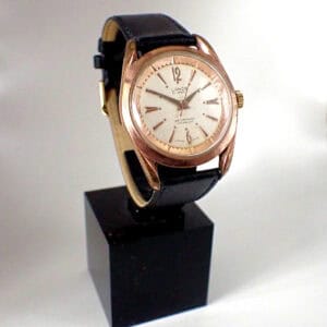 The Lanco 21 Jewels Rare 'Pie Pan' Mechanical features a round gold case, white dial, black leather strap, and minimal hour markers, showcased on a black stand against a white background.