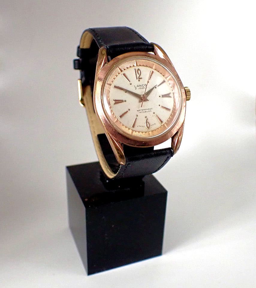 The Lanco 21 Jewels Rare 'Pie Pan' Mechanical features a round gold case, white dial, black leather strap, and minimal hour markers, showcased on a black stand against a white background.