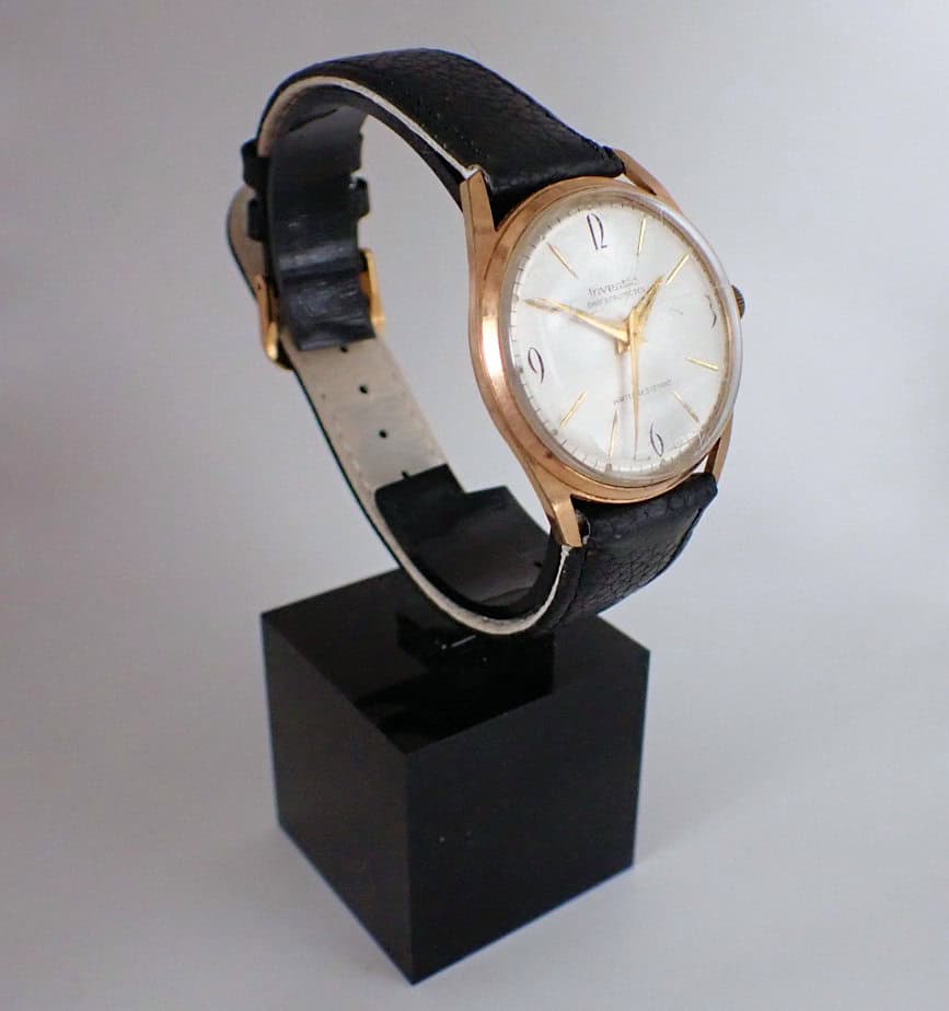 The Inventic Mechanical Circa 1965 - Bargain Basement! features a gold case, white dial, and black leather strap, showcased on a black stand with a plain background.
