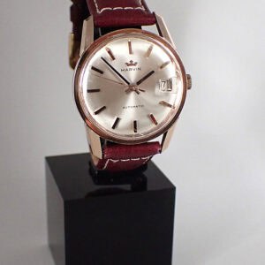 A Marvin Automatic 25 Jewels Cal 776 wristwatch with a round silver dial, gold hour markers, date at 3 o'clock, and brown leather strap is displayed on a black stand against a plain background.