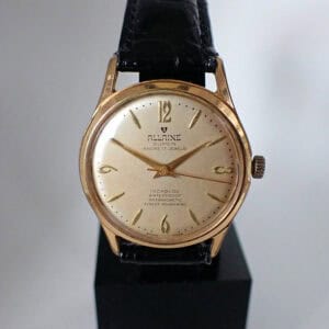 An Allaine Super 'Finest Mainspring' 1950s mechanical wristwatch featuring a round gold case, cream dial with gold hands and numerals, black leather strap, and visible wear on the watch face.