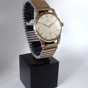 A Certina Mechanical 9ct Gold 1960’s wristwatch with a white dial and metal stretch strap is displayed on a black plinth against a plain, light background.