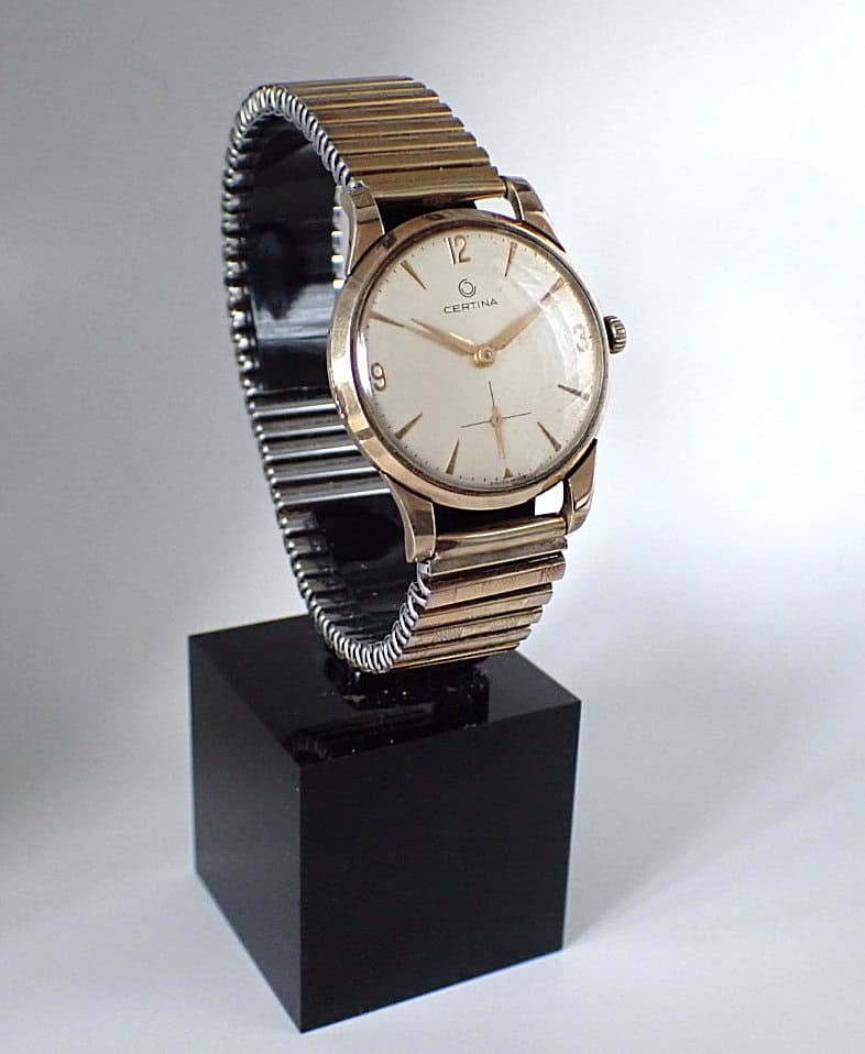 A Certina Mechanical 9ct Gold 1960’s wristwatch with a white dial and metal stretch strap is displayed on a black plinth against a plain, light background.