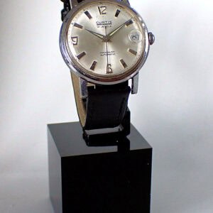 The Curtis Automatic 21 Jewels, a vintage silver wristwatch with a round face and black leather strap, is displayed upright on a black rectangular stand against a plain white background.