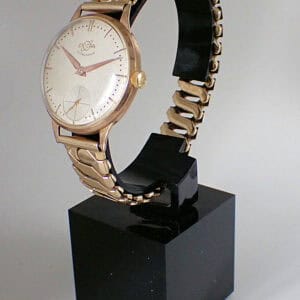 The Enicar Ultrasonic 9ct Gold wristwatch, featuring a round white dial, gold hour markers, and a gold link bracelet, is displayed upright on a black stand against a plain background.