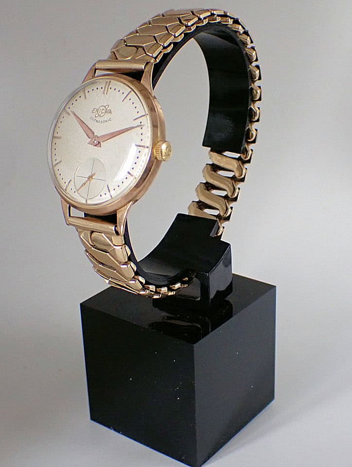 The Enicar Ultrasonic 9ct Gold wristwatch, featuring a round white dial, gold hour markers, and a gold link bracelet, is displayed upright on a black stand against a plain background.