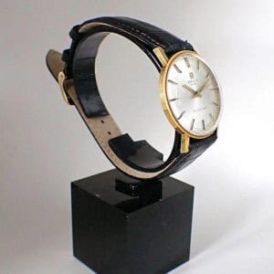 The Tissot Seastar Seven 18ct gold-cased Mechanical 1975 wristwatch with a black leather strap is displayed on a black square stand against a plain white background.