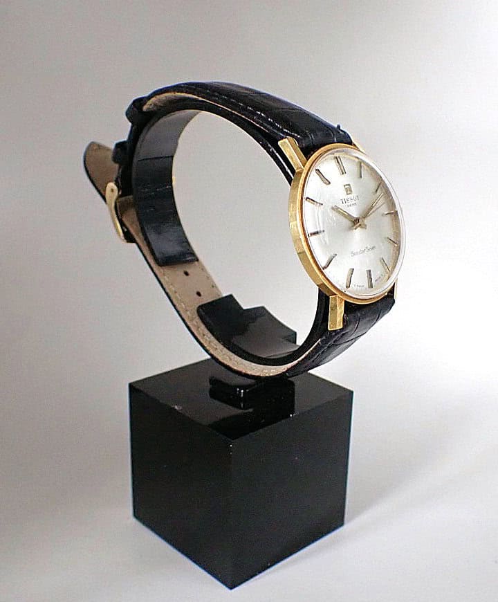 The Tissot Seastar Seven 18ct gold-cased Mechanical 1975 wristwatch with a black leather strap is displayed on a black square stand against a plain white background.