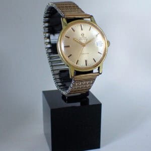 An Omega Gold Plated Genève Automatic 1971 wristwatch with a metal expansion strap, shown upright on a black stand against a plain light background.