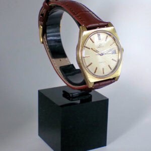 An Omega Gold Plated Genève Mechanical wristwatch with a white dial, date display, and brown leather strap is displayed upright on a black plinth against a neutral background.