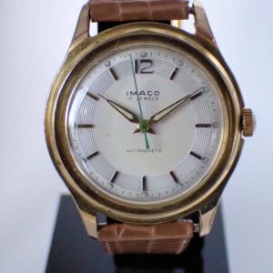 A vintage Imaco 17 Jewels Antimagnetic Mechanical wristwatch with a brown textured leather strap, gold-tone case, and white dial, displayed on a black stand.