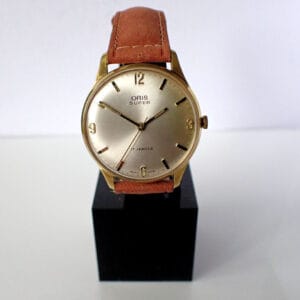 An Oris Super Mechanical Circa 1970 wristwatch with a gold case, beige dial, brown leather strap, and black hour markers is displayed upright on a black stand against a white background.