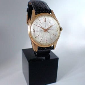 The Benrus Shock Absorber Mechanical 20 Micron Gold Swiss wristwatch, featuring a silver dial and black leather strap, stands upright on a black plinth against a light background.