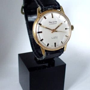 A Paul Jobin of Switzerland Mechanical Swiss Made wristwatch with a gold case, white dial, black leather strap, and gold hour markers is displayed upright on a black stand against a plain background.