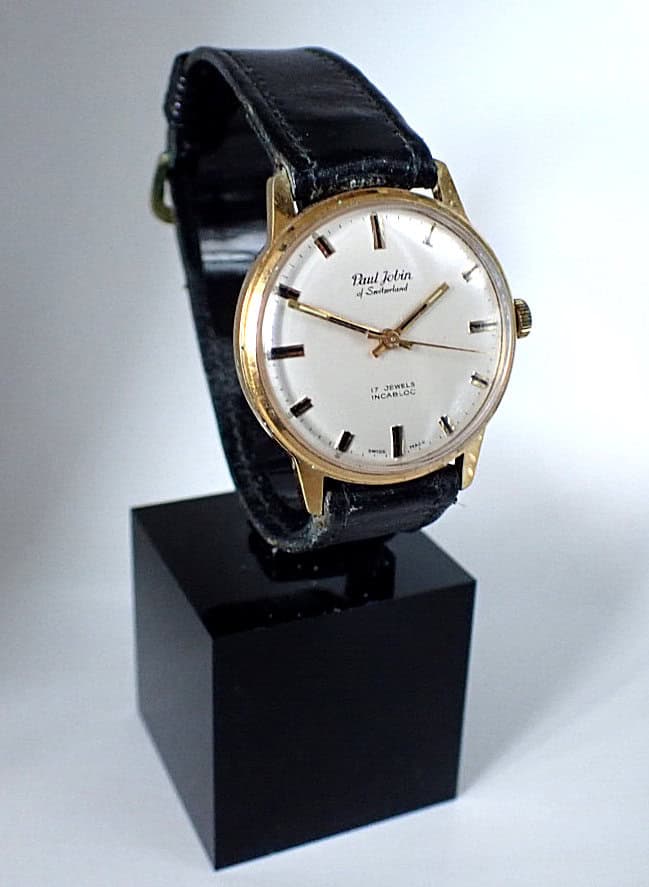 A Paul Jobin of Switzerland Mechanical Swiss Made wristwatch with a gold case, white dial, black leather strap, and gold hour markers is displayed upright on a black stand against a plain background.