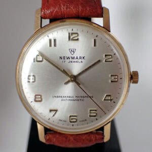 The Newmark Unbreakable Mainspring Mechanical 17 Jewels is a vintage wristwatch with a gold case, brown leather strap, silver dial, and Arabic numerals. The face displays “17 Jewels”, “Unbreakable Mainspring”, and “Antimagnetic”.