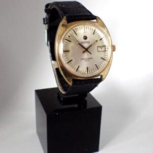 A vintage Roamer Vanguard 303 1960s mechanical wristwatch with a gold case, white dial, black leather strap, and date display is displayed on a black stand against a white background.