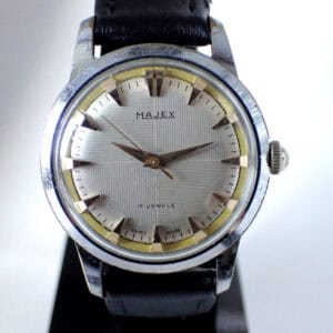 A vintage Majex 17 Jewel Mechanical wristwatch, Swiss made circa 1960s, features a silver dial with gold-tone hour markers and hands, black leather strap, and "17 JEWELS" above 6 o’clock. The watch is upright on a stand.