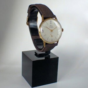 The ELCO Bimatic 15 Jewel Swiss Made Mechanical wristwatch with a brown leather strap, gold case, and white dial with black numerals is displayed on a black stand against a plain light background.