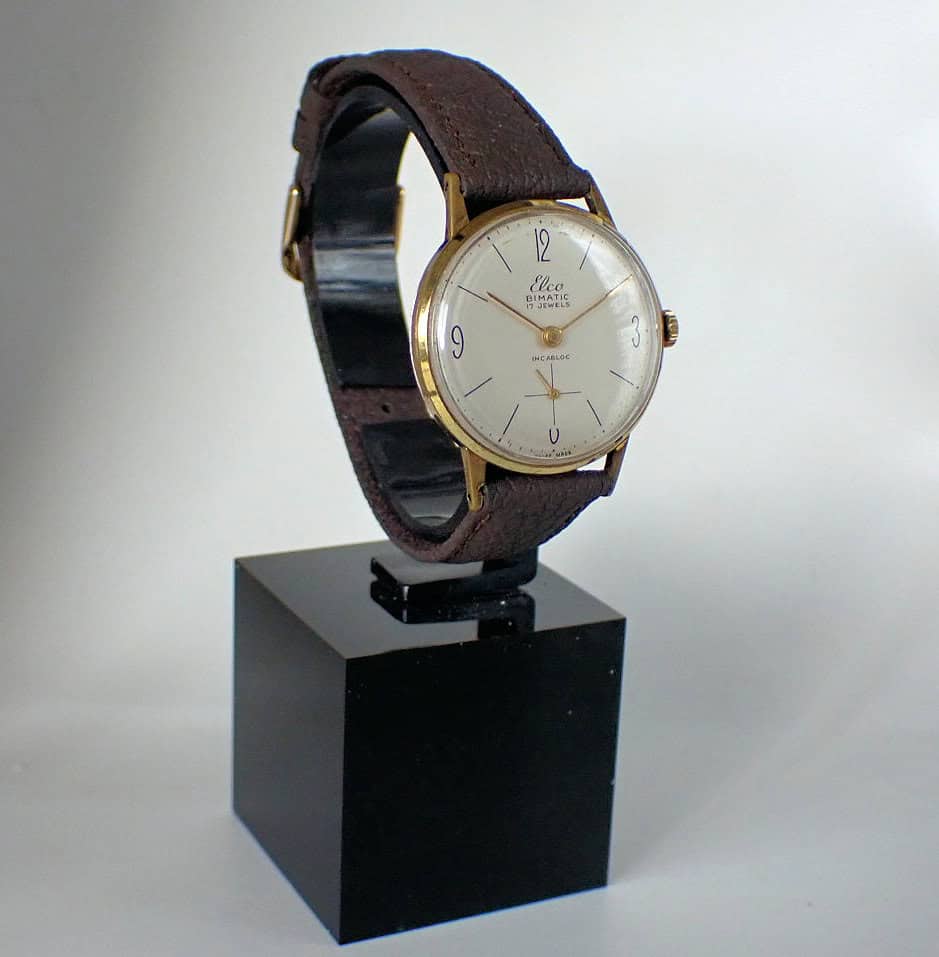 The ELCO Bimatic 15 Jewel Swiss Made Mechanical wristwatch with a brown leather strap, gold case, and white dial with black numerals is displayed on a black stand against a plain light background.